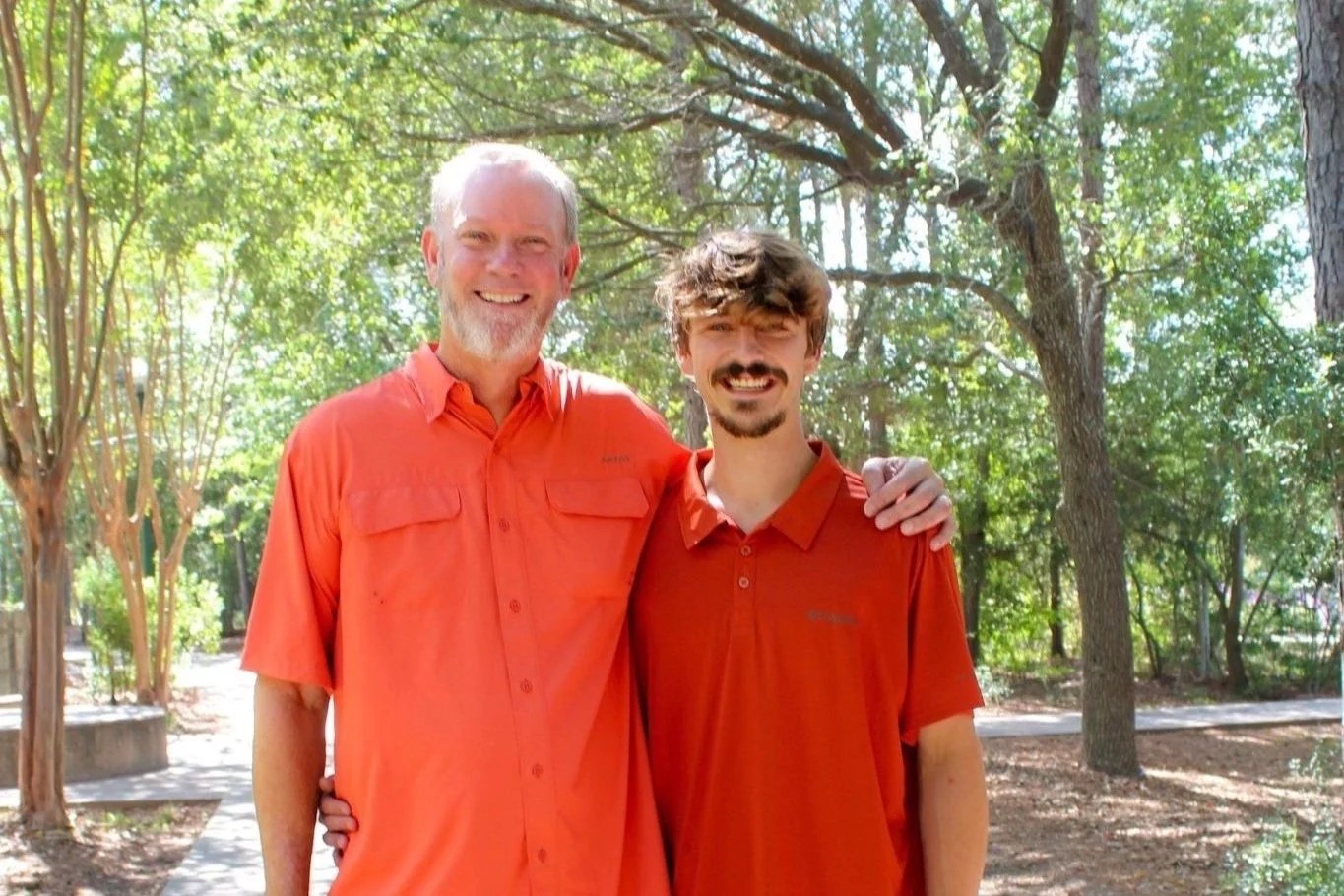 Matt Herron and his son Connor, both in orange shirts, standing under trees